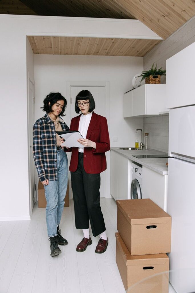 Two women discuss paperwork in a modern kitchen with moving boxes around them, symbolizing a new home purchase.